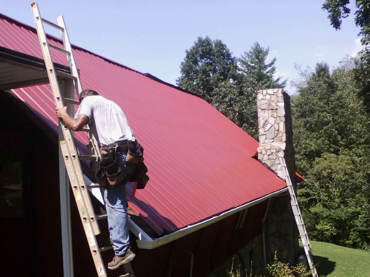 Roofer on a ladder beside a red metal roof and stone chimney — Soffit & Fascia Repair by Roofing Brothers in Thornton